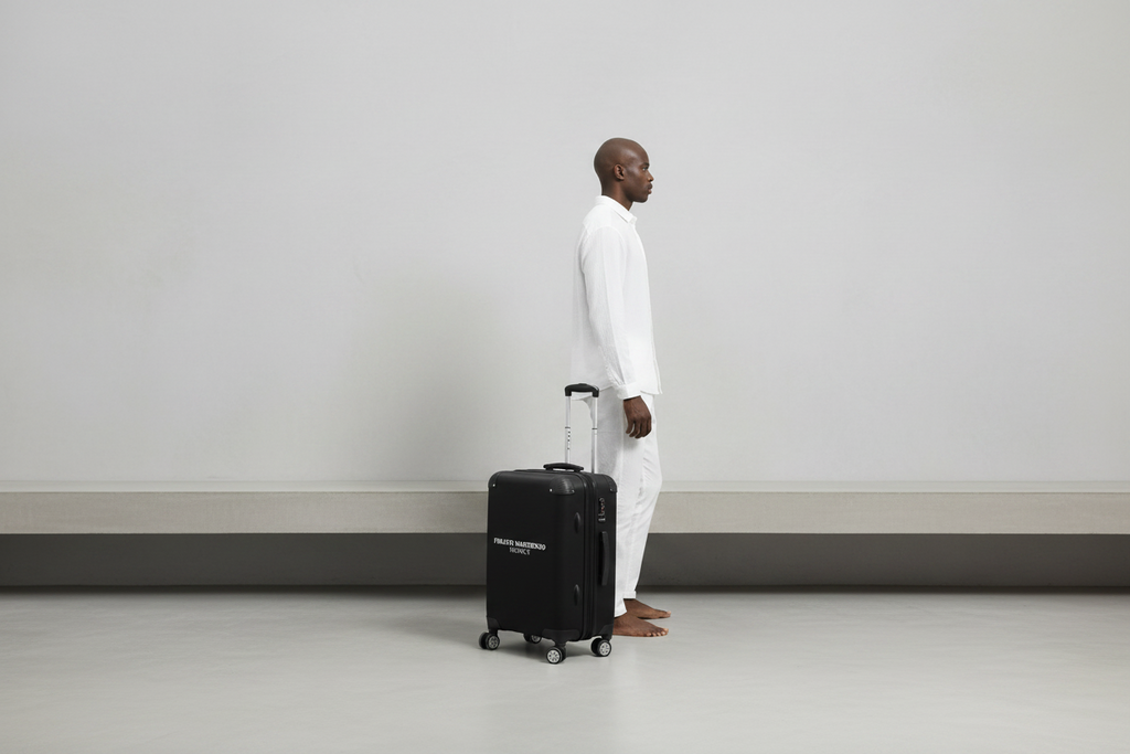 A man in a white shirt and trousers stands in profile next to a black hard-shell suitcase with the brand name 'FREKTI MARTINENO' printed in white on the front, set against a plain white studio background.