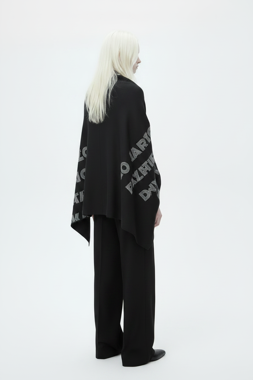 A model with long platinum blonde hair is shown in profile wearing a black shawl with bold white text, paired with black trousers, against a solid white studio background.